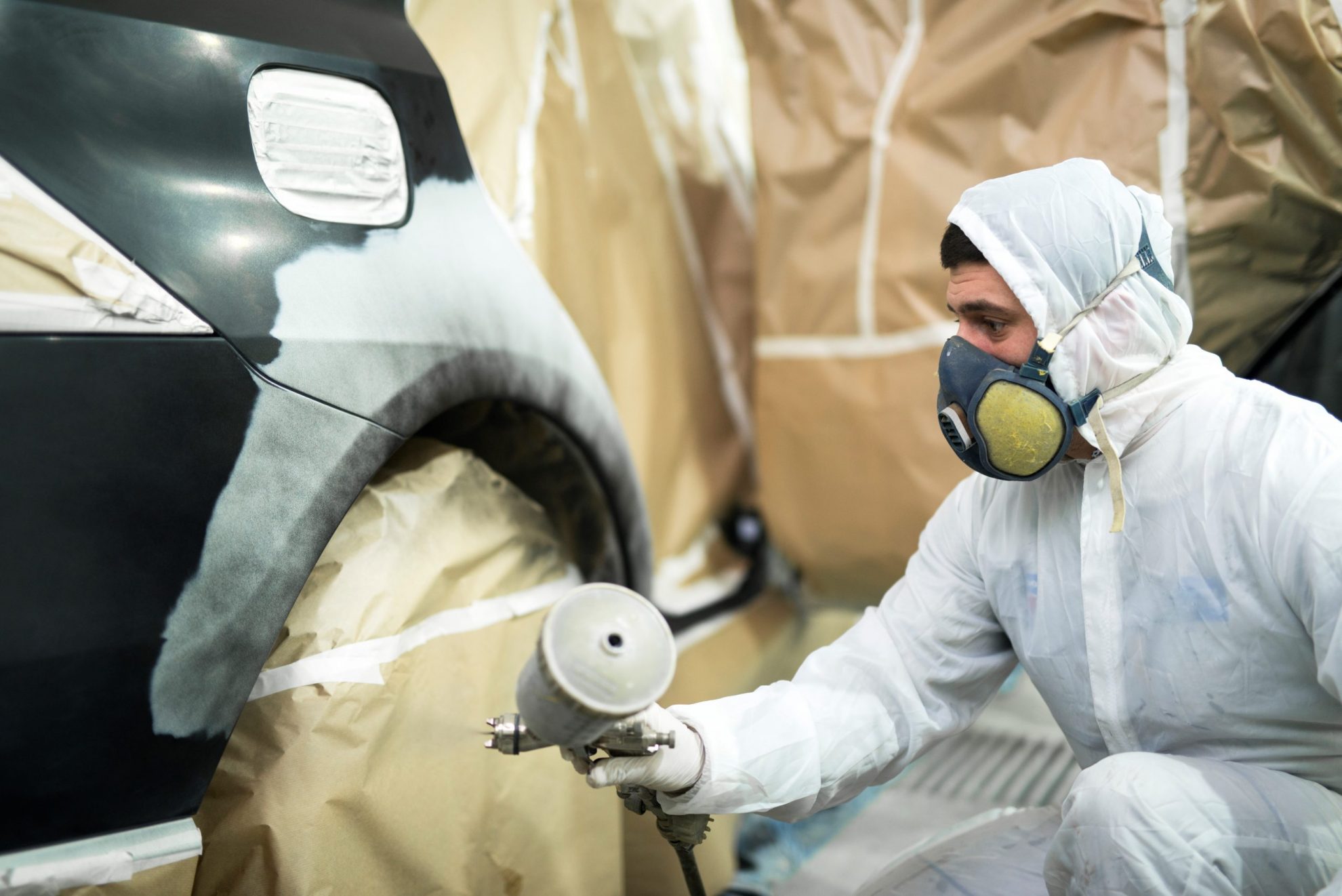 Painter in a protective suit spray-gunning the primed rear quarter panel of a dark car inside a paint booth